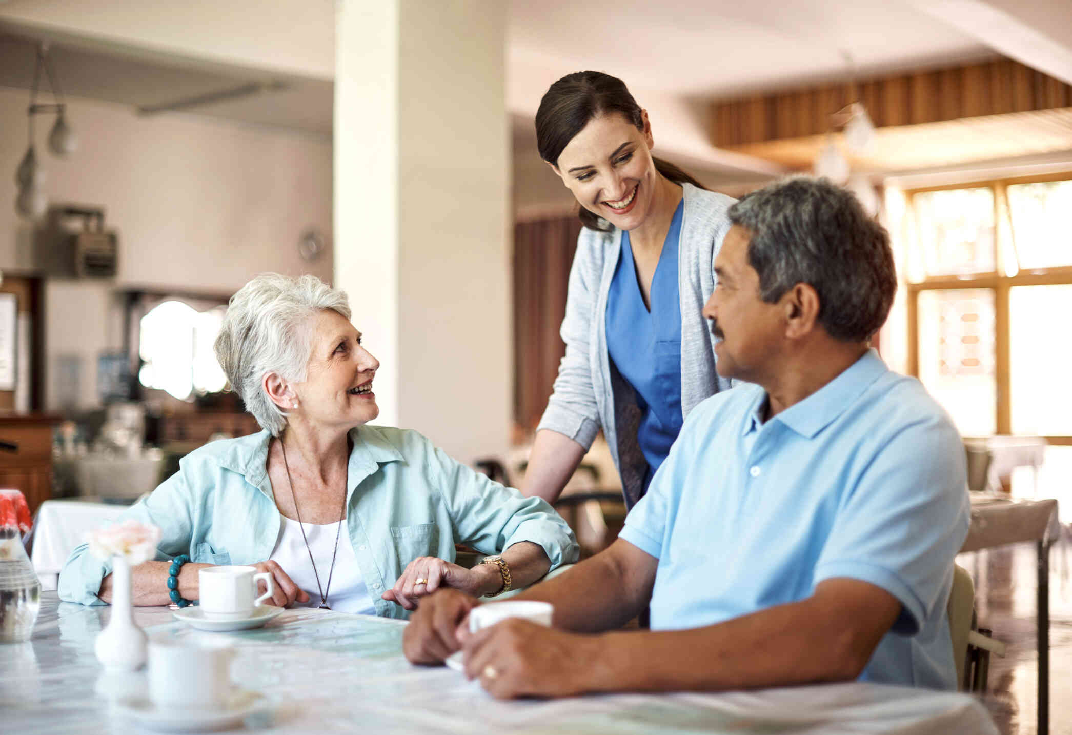 An elderly man and woman sit at a table together as a female nurse comes over to them to check on them while smiling.